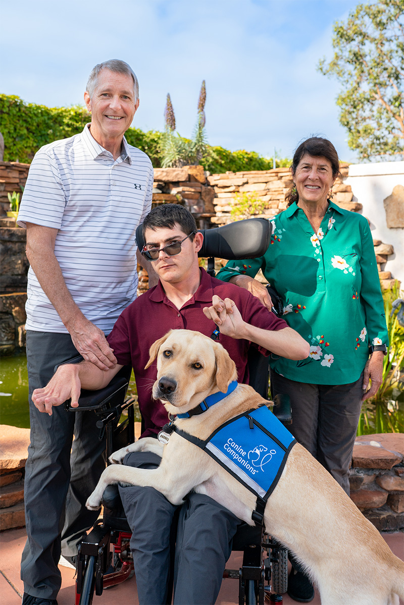 A smiling family with a young man in a power wheelchair with a service dog